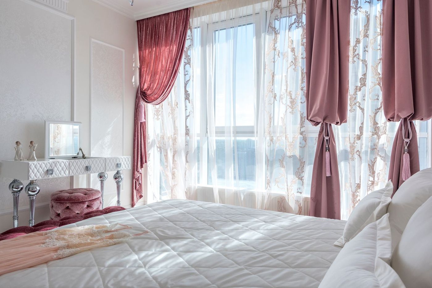 A calm bedroom with pink curtains, tall windows and crisp white bedding